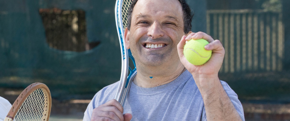 A man smiling. He has a tennis racket over one shoulder and a tennis ball in the other.